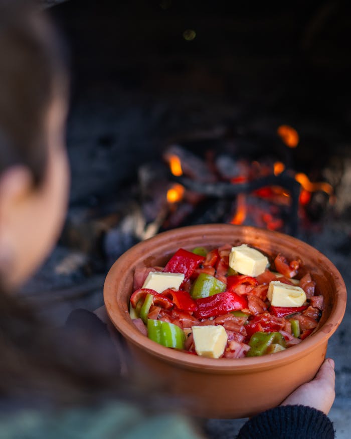 A person holds a clay pot of vegetables and butter near an outdoor fire, ready to cook.