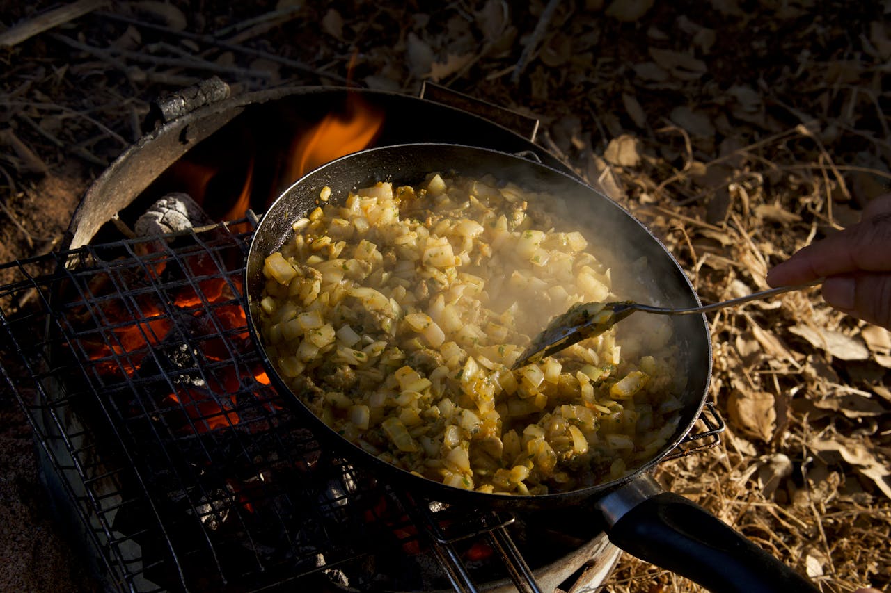 Rustic hash browns cooking on a campfire grill outdoors, emitting steam and aroma.