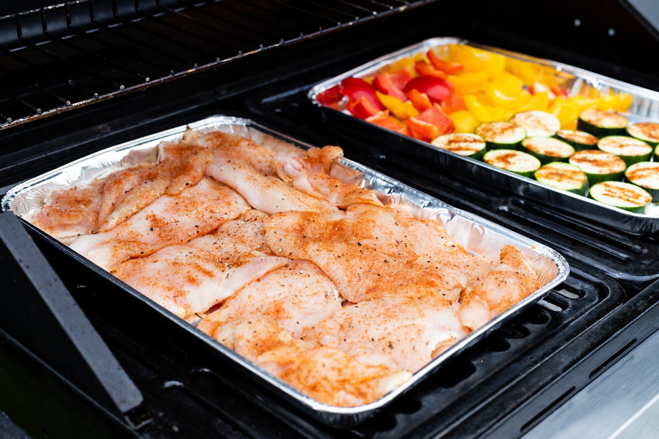 Juicy seasoned chicken and fresh vegetables grilling in aluminum trays for a delicious meal.